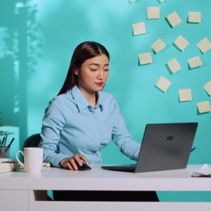 Asian administrative assistant works on a laptop at her desk. Office setting with sticky notes on the wall.
