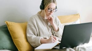 Woman with glasses working on laptop in bed. Headphones, notebook, and pen. Remote work lifestyle.