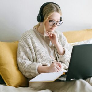 Woman with glasses working on laptop in bed. Headphones, notebook, and pen. Remote work lifestyle.