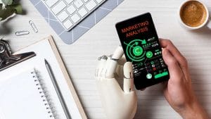 AI for Marketing: Robot hand holding phone with marketing analysis. Keyboard, notepad, and coffee on desk.