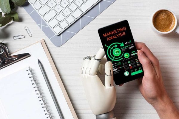 AI for Marketing: Robot hand holding phone with marketing analysis. Keyboard, notepad, and coffee on desk.