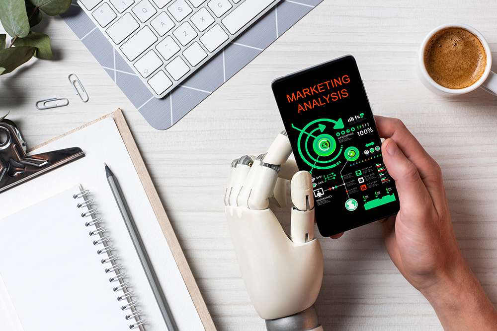 AI for Marketing: Robot hand holding phone with marketing analysis. Keyboard, notepad, and coffee on desk.