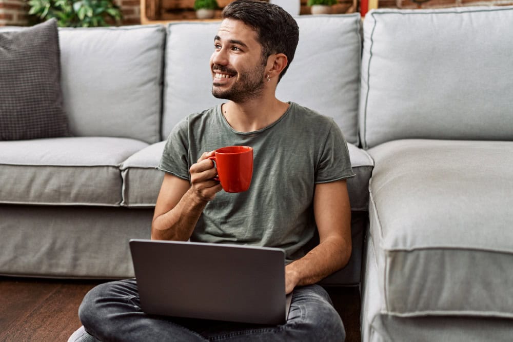 Adult learning mathematics online. Man with laptop and coffee cup smiles, seated on floor.