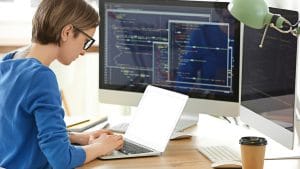 System Security Certified Practitioner (SSCP) at work. Woman coding on laptop with dual monitors.