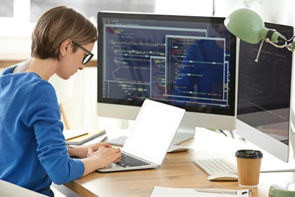 System Security Certified Practitioner (SSCP) at work. Woman coding on laptop with dual monitors.