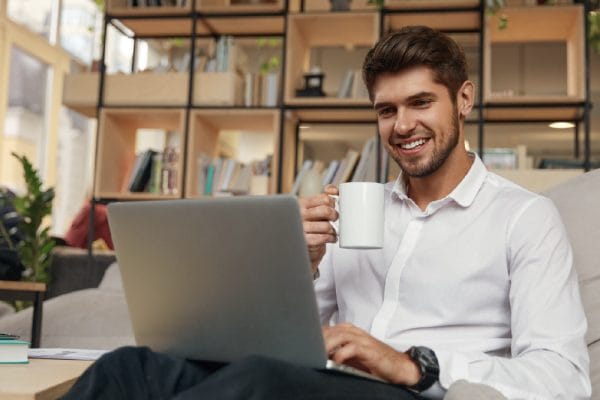Smiling man drinking his morning coffee and working on his laptop.