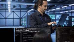 Network admin to site reliability engineer. IT professional with laptop in data center, coding on monitors.