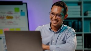 Smiling man using no/low code machine learning on laptop. Whiteboard with sticky notes in background.