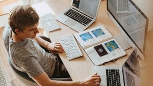 CompTIA Bundle: Man at desk with laptops, keyboard, monitor, and notebook.