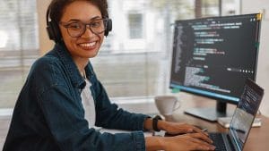 CompTIA Linux+: Woman with headphones codes on laptop. Monitor displays code. Linux+ certification training.
