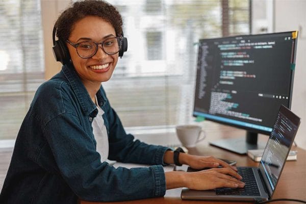 CompTIA Linux+: Woman with headphones codes on laptop. Monitor displays code. Linux+ certification training.