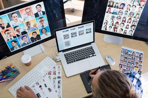 Web design: Designer working with a graphic tablet, laptop, and dual monitors displaying diverse faces.