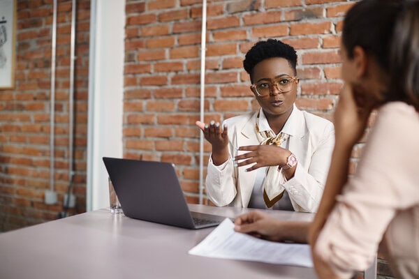 Professional woman in a white blazer and glasses gestures while speaking to a colleague across a desk with a laptop.