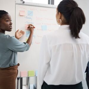 Professional woman with braided hair places a sticky note on a whiteboard while two colleagues observe in an office.
